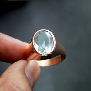 Puede incluir: Un anillo dorado con una gema ovalada y transparente. El anillo tiene una banda gruesa y pulida, y la gema est&aacute; engastada en un bisel. El anillo est&aacute; sostenido por los dedos de una persona.