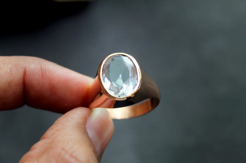 Puede incluir: Un anillo dorado con una gema ovalada y transparente. El anillo tiene una banda gruesa y pulida, y la gema est&aacute; engastada en un bisel. El anillo est&aacute; sostenido por los dedos de una persona.