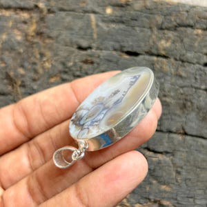 May include: Oval pendant with a silver bezel. The pendant features a natural stone with a unique pattern of beige, white, and grey hues. The stone has a polished surface and a silver loop for attaching to a chain.