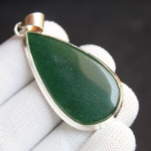 May include: A teardrop-shaped pendant with a polished, deep green stone set in a silver-colored metal frame. The pendant has a small, gold-colored bail for a chain. The background is dark.