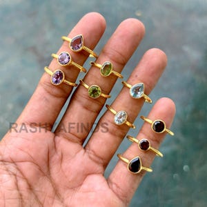 May include: A collection of gold-colored rings with various gemstone settings, including amethyst, peridot, aquamarine, and garnet. The gemstones are oval, pear, and round-cut. The rings are displayed on a hand. The text "RASHYAFINDS" is visible.