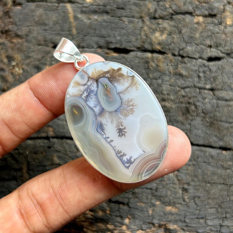 May include: Oval pendant with a polished agate stone. The stone has a natural landscape pattern in white, grey, brown, and green. It is set in a silver-coloured metal bezel with a loop for a chain.