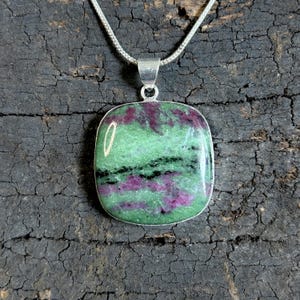 Peut inclure: Un collier avec un pendentif carré orné d'une pierre précieuse verte et rose sertie dans un cadre en métal argenté. La pierre a un aspect marbré avec des inclusions vertes foncées, roses et noires. La chaîne est argentée.