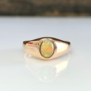 Könnte beinhalten: Ein roségoldener Ring mit einem ovalen Opal-Edelstein. Der Opal hat gelbe, orange und grüne Farbtöne. Der Ring liegt auf einer weißen Oberfläche.