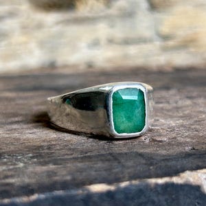 Può includere: Un anello d'argento con una grande gemma di smeraldo rettangolare. Lo smeraldo è di un colore verde vibrante ed è incastonato in una fascia d'argento lucida. L'anello è esposto su una superficie di legno.