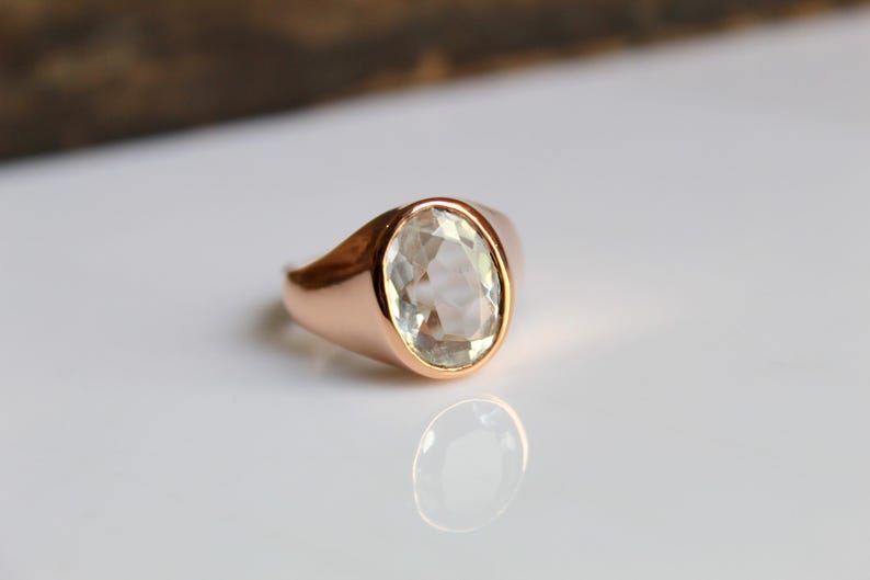 Puede incluir: Un anillo de oro rosa con una gema grande, de talla ovalada, de color verde p&aacute;lido. El anillo est&aacute; engastado en una banda lisa y pulida, y la gema refleja la luz. El anillo se exhibe sobre una superficie blanca.