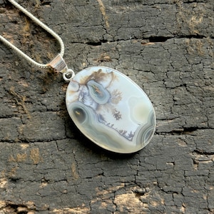May include: Oval pendant necklace with a polished agate stone. The stone has a unique pattern in white, grey, and brown. It's set in silver-tone metal and hangs from a delicate silver chain. The necklace is a stylish accessory.