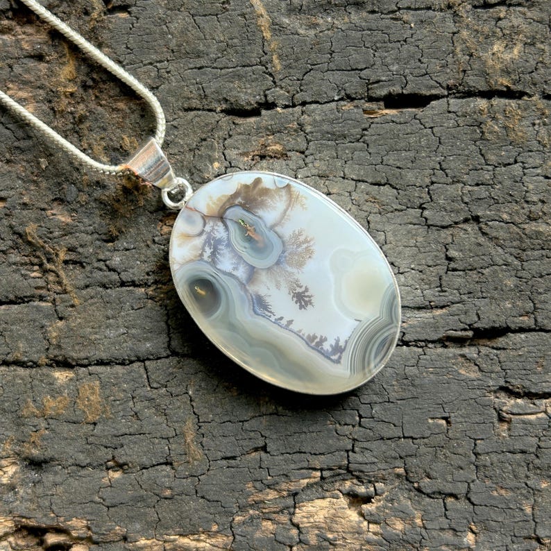 May include: Oval pendant necklace with a polished agate stone. The stone has a unique pattern in white, grey, and brown. It's set in silver-tone metal and hangs from a delicate silver chain. The necklace is a stylish accessory.
