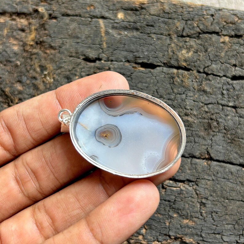 May include: Oval pendant with a polished agate stone set in a silver-coloured metal frame. The stone shows concentric circles in white, brown, and tan. The pendant has a loop for attaching to a chain. The stone's pattern resembles an eye.
