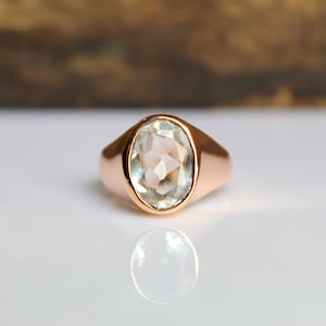 Puede incluir: Un anillo de oro rosa con una piedra preciosa ovalada, facetada y de color claro. El anillo est&aacute; sobre una superficie blanca con un fondo marr&oacute;n borroso. La piedra preciosa refleja la luz y la banda del anillo tiene un acabado liso y pulido.