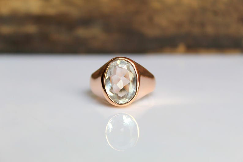 Puede incluir: Un anillo de oro rosa con una piedra preciosa ovalada, facetada y de color claro. El anillo est&aacute; sobre una superficie blanca con un fondo marr&oacute;n borroso. La piedra preciosa refleja la luz y la banda del anillo tiene un acabado liso y pulido.