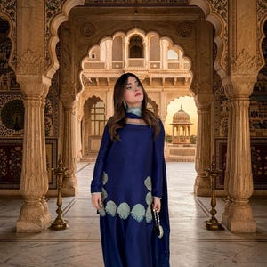 May include: A navy blue ensemble featuring a long-sleeved top with decorative floral embroidery and a matching flowing skirt. The outfit includes a light blue scarf and is accessorized with a beaded clutch. The background showcases an ornate architectural setting.
