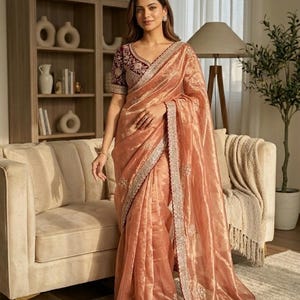 Puede incluir: Un sari color melocotón con intrincados bordados plateados en los bordes. El sari se usa con una blusa burdeos a juego con detalles. La tela tiene una calidad brillante y translúcida, creando una apariencia elegante y fluida.