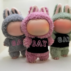 May include: Three plush dolls in bunny costumes. The dolls have large, round faces with rosy cheeks and wear fuzzy outfits. One doll is pink with a black shirt that says "GAY", the others are gray and green with "GA" on their shirts.