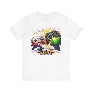 Op de afbeelding: Wit t-shirt met een kleurrijke afbeelding van twee gamecontrollers die aan het boksen zijn, met de tekst "BUTTON SMASH BATTLE". De controllers zijn afgebeeld met bokshandschoenen en in een dynamische pose.