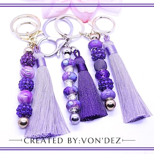 May include: Keychains with purple beaded accents and tassels. The word "LOVE" is displayed vertically on the left side. The keychains have silver-colored hardware and are labeled "CREATED BY: VON'DEZ".