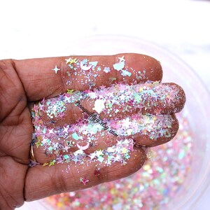 May include: A close-up of a hand holding iridescent glitter with a mix of star, crescent moon, and other shapes. The glitter is in a variety of colors, including pink, blue, green, and white.