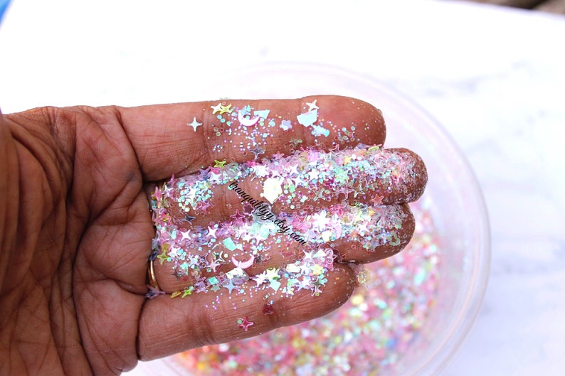 May include: A close-up of a hand holding iridescent glitter with a mix of star, crescent moon, and other shapes. The glitter is in a variety of colors, including pink, blue, green, and white.