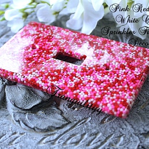 Könnte beinhalten: Ein Lichtschalter-Abdeckung mit rosa, roten und weißen Zuckerstreuseln, die in klarem Harz eingebettet sind. Die Abdeckung befindet sich auf einer grauen, strukturierten Oberfläche.