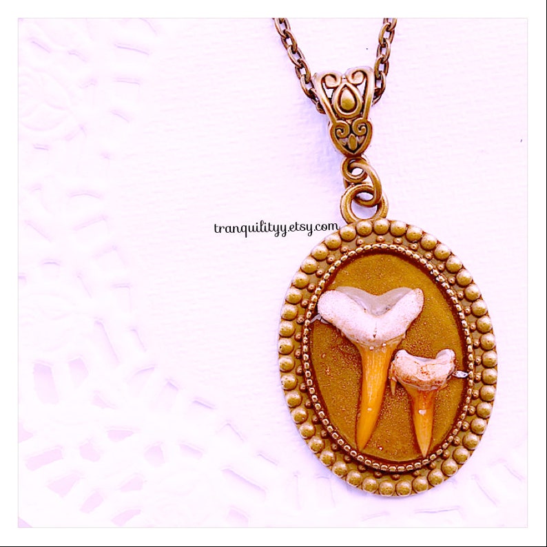 May include: A gold pendant necklace with a round, ornate frame. Inside the frame are two fossilized shark teeth, one larger than the other. The teeth are white and have a natural, organic texture. The pendant is hanging from a gold chain.