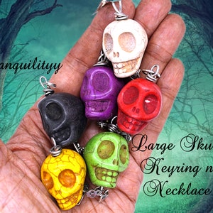 May include: A collection of six colorful skull charms, each with a different color and design. The charms are made of stone and have a smooth, polished finish. The charms are strung on a silver chain and are perfect for making a necklace or keychain.  "Large Skull Keyring n Necklace" is written in cursive on the image.