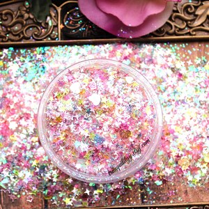 May include: A clear jar filled with iridescent pink, white, and blue glitter in the shape of stars and hearts. The glitter is on a wooden surface with a gold ornate frame.