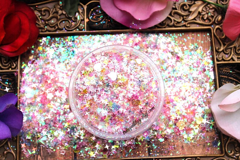 May include: A clear jar filled with iridescent pink, white, and blue glitter in the shape of stars and hearts. The glitter is on a wooden surface with a gold ornate frame.