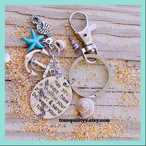 Puede incluir: Un llavero de plata con un dije que dice "May you always have a shell in your pocket & sand between your toes". El dije está decorado con una estrella de mar azul, un ancla plateada y un caballito de mar plateado. El llavero está sobre una superficie de madera con arena.