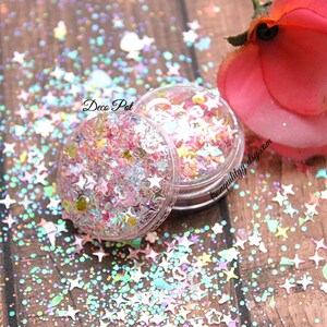 May include: A small clear jar filled with iridescent pink, blue, and white glitter in the shape of stars and circles. The jar is labeled "Deco Pot" and is surrounded by loose glitter on a wooden surface.