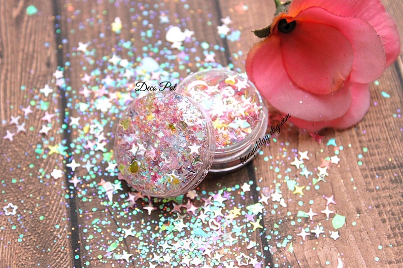 May include: A small clear jar filled with iridescent pink, blue, and white glitter in the shape of stars and circles. The jar is labeled "Deco Pot" and is surrounded by loose glitter on a wooden surface.
