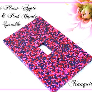 Könnte beinhalten: Eine rechteckige Lichtschalterabdeckung, verziert mit roten, rosa und lila Streuseln. Der Text "Sweet Plums, Apple, Red & Pink Candy, Sprinkle" ist kursiv geschrieben. Das Wort "Tranquilityy" ist rechts unten kursiv geschrieben.