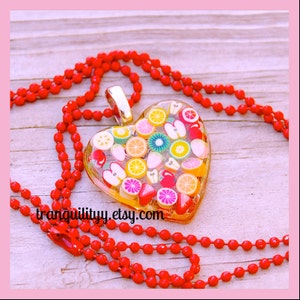 Puede incluir: Un collar de cuentas rojas con un colgante en forma de corazón. El colgante está lleno de rodajas de fruta de colores, incluyendo kiwi, manzana y cítricos.