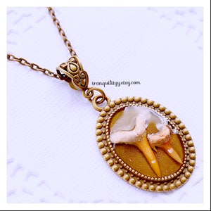 May include: A gold oval pendant necklace with a clear resin setting featuring two fossilized shark teeth. The pendant is surrounded by a decorative border of small beads. The necklace is on a chain with a decorative clasp. The text "tranquilityy.etsy.com" is visible in the image.