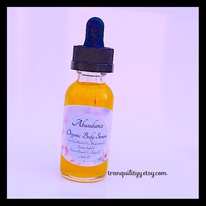 Pu&ograve; includere: Una bottiglia di vetro trasparente riempita con un liquido giallo dorato, etichettata "Abundance Organic Body Serum". La bottiglia ha un contagocce nero e un'etichetta bianca con decorazioni floreali. L'URL del negozio Etsy "tranquilityy.etsy.com" &egrave; visibile.