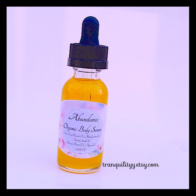 Pu&ograve; includere: Una bottiglia di vetro trasparente riempita con un liquido giallo dorato, etichettata "Abundance Organic Body Serum". La bottiglia ha un contagocce nero e un'etichetta bianca con decorazioni floreali. L'URL del negozio Etsy "tranquilityy.etsy.com" &egrave; visibile.