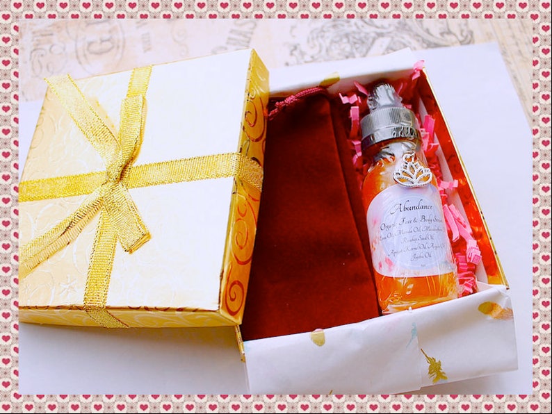 Pu&ograve; includere: Confezione regalo bianca con fiocco dorato, contenente un flacone di siero biologico per viso e corpo "Abundance". La bottiglia &egrave; circondata da carta rosa e un sacchetto di velluto rosso. La scatola ha un motivo floreale dorato.