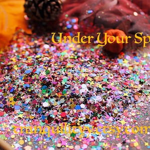 May include: A pile of colorful glitter with stars and other shapes, on a wooden surface. The text "Under Your Spell" is written in yellow above the glitter. The text "tranquilityetsy.com" is written in yellow below the glitter.