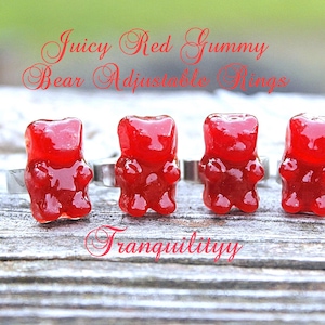 Op de afbeelding: Vier rode gummy bear ringen op verstelbare zilveren bandjes. De ringen zijn in een rij op een houten oppervlak gerangschikt. De tekst "Juicy Red Gummy Bear Adjustable Rings" staat boven de ringen en de tekst "Tranquilityy" onder de ringen.