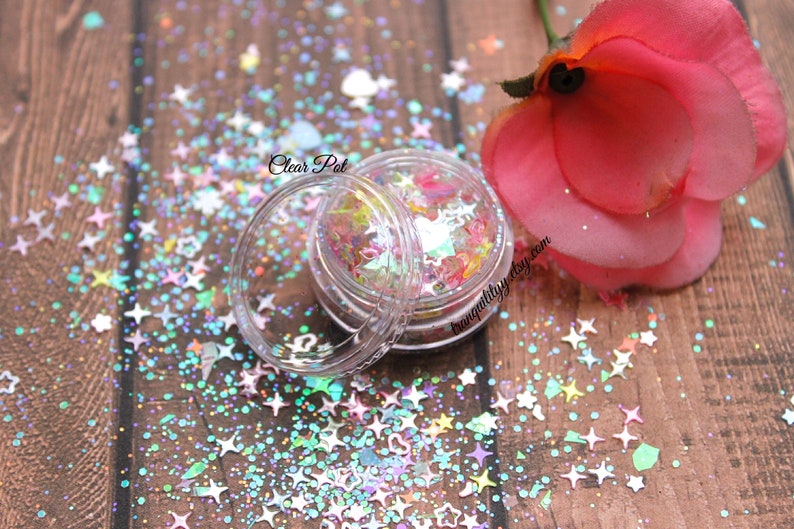 May include: A clear plastic jar filled with iridescent star-shaped glitter. The glitter is a mix of pastel colors, including pink, blue, green, and yellow. The jar is sitting on a wooden surface with a pink rose in the background. The text "Clear Pot" is visible on the jar.