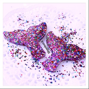 Puede incluir: Dos encantos de cola de sirena brillantes hechos de resina y llenos de purpurina colorida. Las colas son de color morado claro con una variedad de colores de purpurina, incluyendo rosa, azul, verde y dorado.