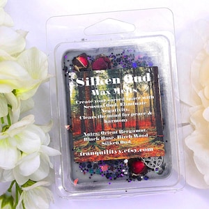 May include: A clear plastic container holding a dark gray wax melt with decorative glitter and dried flowers. The label reads "Silken Oud Wax Melts" with scent notes listed. The background features white flowers.