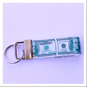 May include: A white key chain with a green and white design of a $100 bill. The key chain has a silver key ring and a gold metal clasp. The text on the bill reads "United States of America" and "FE 523399545 A".