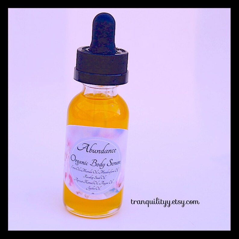 Pu&ograve; includere: Una bottiglia di vetro trasparente riempita con un liquido dorato, etichettata "Abundance Organic Body Serum". L'etichetta elenca vari oli. La bottiglia ha un contagocce nero. Il sito web "tranquilityy.etsy.com" &egrave; stampato in basso a destra.