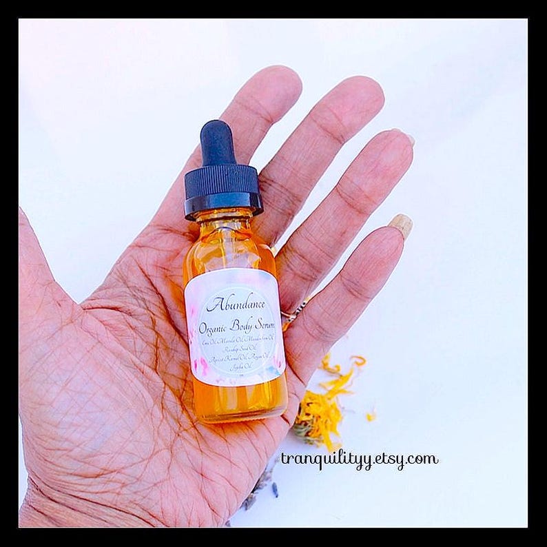 Pu&ograve; includere: Una piccola bottiglia di vetro ambrato con un contagocce nero, etichettata "Abundance Organic Body Serum". La bottiglia &egrave; tenuta in una mano aperta. Lo sfondo &egrave; chiaro con alcuni petali di fiori e l'indirizzo del sito web tranquilityyy.etsy.com.
