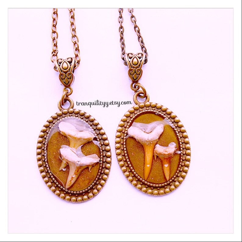 May include: Two bronze oval pendants with a gold-toned chain. Each pendant features a clear resin oval with three fossilized shark teeth inside. The text "tranquilityy.etsy.com" is printed on the bottom of the image.