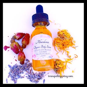 Pu&ograve; includere: Una bottiglia di vetro trasparente di "Abundance Organic Body Serum" con un contagocce blu, circondata da fiori secchi, boccioli di rosa e lavanda. Il siero &egrave; color ambra e l'etichetta elenca vari oli. Il sito web "tranquilityyetsy.com" &egrave; visibile.