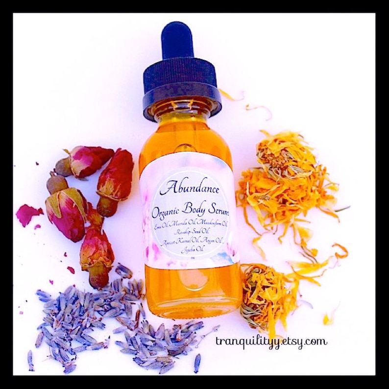 Pu&ograve; includere: Una bottiglia di vetro trasparente di "Abundance Organic Body Serum" con un contagocce blu, circondata da fiori secchi, boccioli di rosa e lavanda. Il siero &egrave; color ambra e l'etichetta elenca vari oli. Il sito web "tranquilityyetsy.com" &egrave; visibile.
