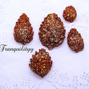 Op de afbeelding: Vijf bruine en gouden glitter dennenappelvormige ornamenten. De ornamenten liggen op een wit kantonderzetter.