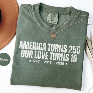 May include: A moss green short-sleeve t-shirt with white text that reads: AMERICA TURNS 250 OUR LOVE TURNS 10, 1776 - 2016 - 2026. The shirt features a Comfort Colors brand label at the inner neckline indicating size S, P, CH.