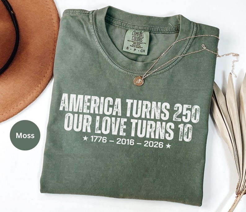 May include: A moss green short-sleeve t-shirt with white text that reads: AMERICA TURNS 250 OUR LOVE TURNS 10, 1776 - 2016 - 2026. The shirt features a Comfort Colors brand label at the inner neckline indicating size S, P, CH.